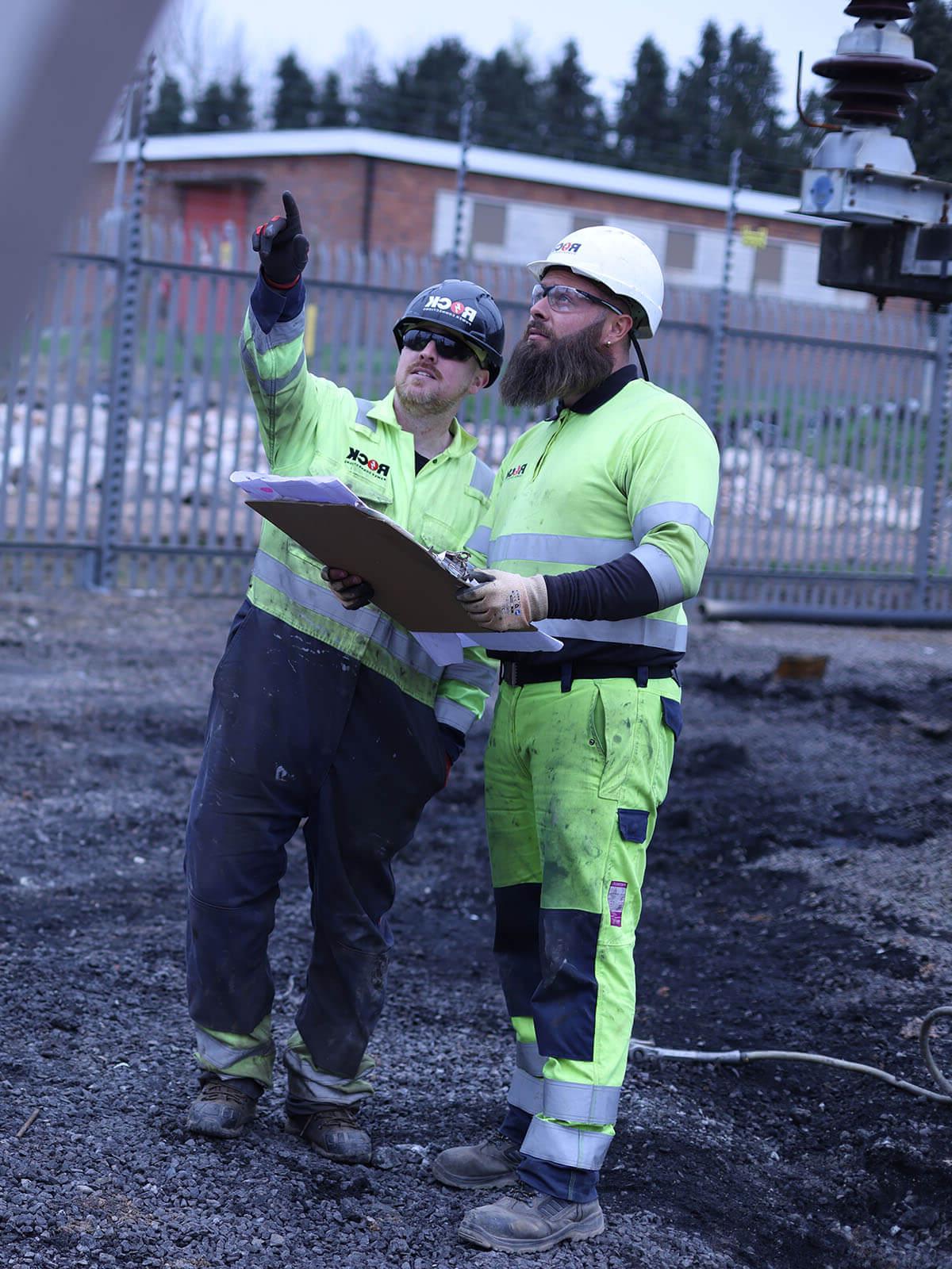 Two male Rock Power Connections employees in high vis clothing, looking at plans and pointing upwards