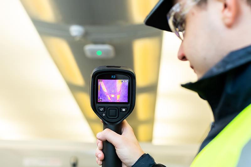 A man in protective equipment, including plastic glasses and high-vis jacket, uses a handheld thermal camera at building vents