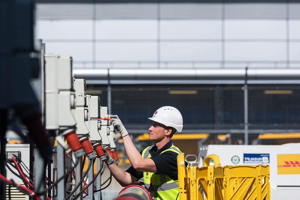 An engineer using a screwdriver to fix an electrical charge point outside of London Heathrow Airport. He is wearing a Mitie-branded hard hat and high vis vest, with yellow safety barriers behind him